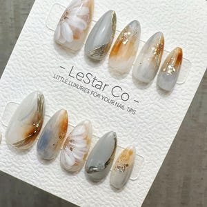 May include: A set of almond-shaped press-on nails. The nails feature a variety of designs, including white floral patterns, gold accents, and glitter. The color palette includes white, gray, and orange tones. The text "- LeStar Co - LITTLE LUXURIES FOR YOUR NAIL TIPS" is visible.