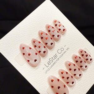 May include: A set of almond-shaped, press-on nail tips. The nails are a light pink with a glittery finish and small red heart accents. The text "- LeStar Co - LITTLE LUXURIES FOR YOUR NAIL TIPS" is printed on the packaging.