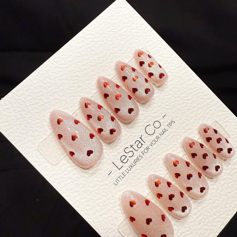 May include: A set of almond-shaped, press-on nail tips. The nails are a light pink with a glittery finish and small red heart accents. The text "- LeStar Co - LITTLE LUXURIES FOR YOUR NAIL TIPS" is printed on the packaging.
