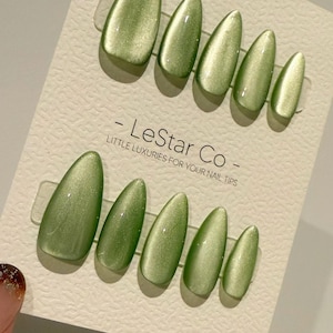 Puede incluir: Un juego de uñas postizas verde oliva de varios tamaños, dispuestas sobre una tarjeta beige claro. Las uñas tienen un acabado brillante y forma ovalada. La tarjeta tiene el texto "- LeStar Co - LITTLE LUXURIES FOR YOUR NAIL TIPS."