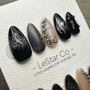 Könnte beinhalten: Ein Set ovaler Kunstnägel in verschiedenen Designs. Die Nägel sind in Schwarz, Silberglitter und Nude gehalten, mit silbernen Verzierungen und abstrakten Mustern. Der Text "- LeStar Co - LITTLE LUXURIES FOR YOUR NAIL TIPS" ist sichtbar.