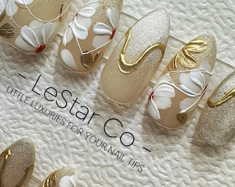 May include: Close-up of a set of artificial nails with a floral design. The nails are almond-shaped with a nude base, white flowers, gold accents, and glitter. The text "- Lestar Co - LITTLE LUXURIES FOR YOUR NAIL TIPS" is visible.