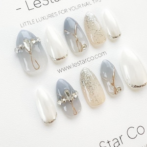 Pode incluir: Conjunto de dez unhas posti&ccedil;as com um design degrad&ecirc; em cinza e branco. Algumas unhas t&ecirc;m glitter prateado, pedrarias e detalhes dourados. As unhas t&ecirc;m formato de am&ecirc;ndoa. A imagem inclui o texto "LeStar Co - LITTLE LUXURIES FOR YOUR NAIL TIPS" e "www.lestarco.com".