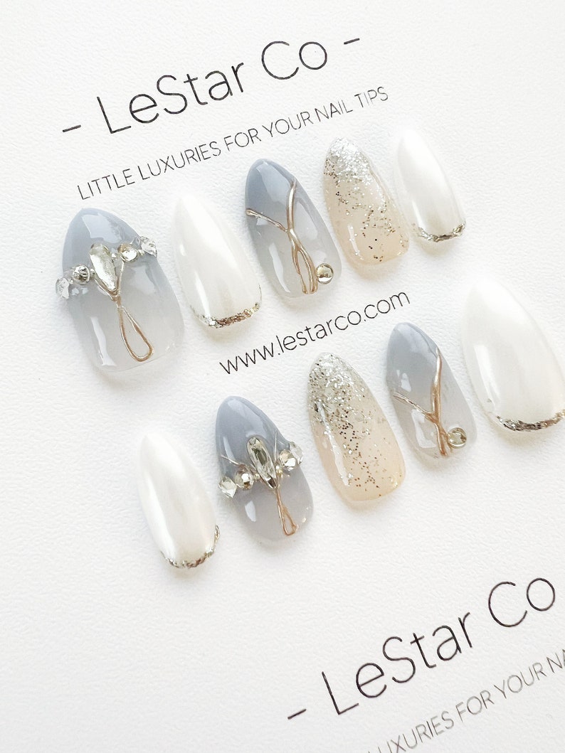Pode incluir: Conjunto de dez unhas posti&ccedil;as com um design degrad&ecirc; em cinza e branco. Algumas unhas t&ecirc;m glitter prateado, pedrarias e detalhes dourados. As unhas t&ecirc;m formato de am&ecirc;ndoa. A imagem inclui o texto "LeStar Co - LITTLE LUXURIES FOR YOUR NAIL TIPS" e "www.lestarco.com".