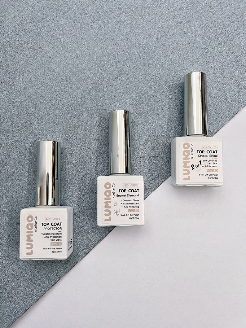 May include: Three bottles of clear nail polish with silver caps. Each bottle has a white label with the brand name "LUMIGO" and the text "NO WIPE TOP COAT" followed by the type of top coat: "PROTECTOR", "Enamel Diamond", and "Crystal Shine".