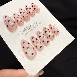 May include: Set of almond-shaped false nails with a glittery pink base and small red heart designs. The nails are arranged on a white card with the text "- LeStar Co - LITTLE LUXURIES FOR YOUR NAIL TIPS".