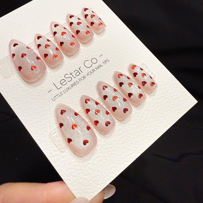 May include: Set of almond-shaped false nails with a glittery pink base and small red heart designs. The nails are arranged on a white card with the text "- LeStar Co - LITTLE LUXURIES FOR YOUR NAIL TIPS".