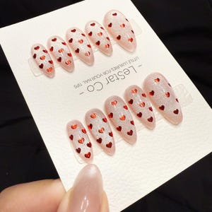 May include: Set of almond-shaped false nails with a glittery pink base and small red heart designs. The nails are arranged on a white card with the text "- LESTAR CO - LITTLE LUXURIES FOR YOUR NAIL TIPS".