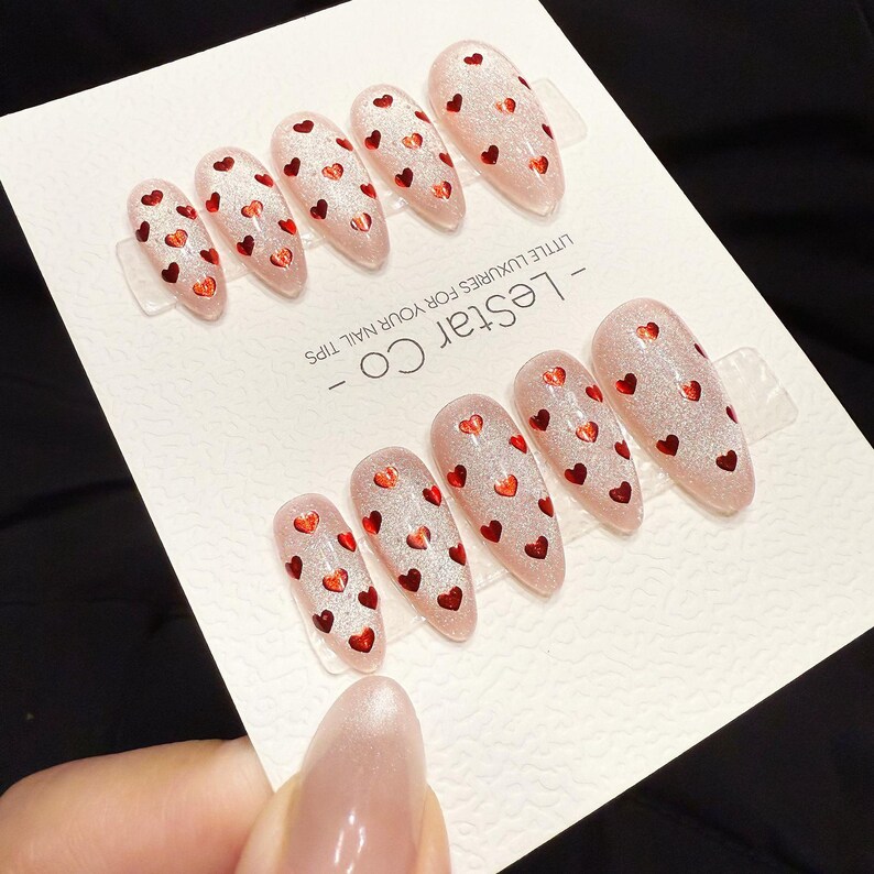 May include: Set of almond-shaped false nails with a glittery pink base and small red heart designs. The nails are arranged on a white card with the text "- LESTAR CO - LITTLE LUXURIES FOR YOUR NAIL TIPS".