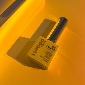 May include: A yellow bottle of Lumigo Gel Colour nail polish with the text "Gel Colour Sheer Colour" and a list of features: Long Wear, Smooth Apply, Fast Cure, Soak-Off Gel Polish. The bottle is on a yellow surface.