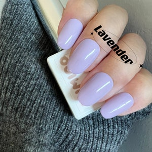 May include: A hand model wearing lavender nail polish. The polish is a light purple shade with a glossy finish. The word "Lavender" is written in black text on a white circle.