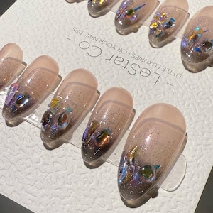 Puede incluir: Un conjunto de uñas postizas con un diseño degradado, que va del rosa claro a un tono más oscuro. Cada uña está adornada con purpurina iridiscente y adornos coloridos de forma irregular. El texto "- LeStar Co - LITTLE LUXURIES FOR YOUR NAIL TIPS" es visible.