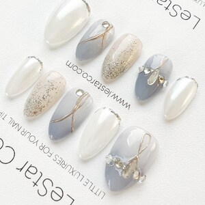 Pode incluir: Um conjunto de dez unhas posti&ccedil;as com um design cinza claro, branco e glitter dourado. As unhas t&ecirc;m formato de am&ecirc;ndoa e um acabamento brilhante. O texto "LeStar Co - LITTLE LUXURIES FOR YOUR NAIL" e "www.lestarco.com" est&aacute; impresso no fundo.