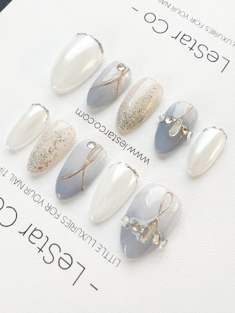 Pode incluir: Um conjunto de dez unhas posti&ccedil;as com um design cinza claro, branco e glitter dourado. As unhas t&ecirc;m formato de am&ecirc;ndoa e um acabamento brilhante. O texto "LeStar Co - LITTLE LUXURIES FOR YOUR NAIL" e "www.lestarco.com" est&aacute; impresso no fundo.