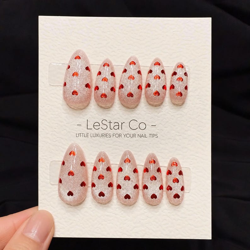 May include: A set of almond-shaped false nails with a glittery pink base and small red heart accents. The nails are arranged in two rows on a white card, with the text "- LeStar Co - LITTLE LUXURIES FOR YOUR NAIL TIPS".