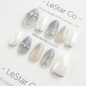 Pode incluir: Conjunto de dez unhas posti&ccedil;as com um design branco, cinza e prata com glitter. As unhas apresentam um detalhe de glitter prata, um design de espiral prata e um strass prata. As unhas est&atilde;o dispostas sobre um fundo branco com o texto "LeStar Co - Little Luxuries for Your Nail Tips" e "www.lestarco.com".