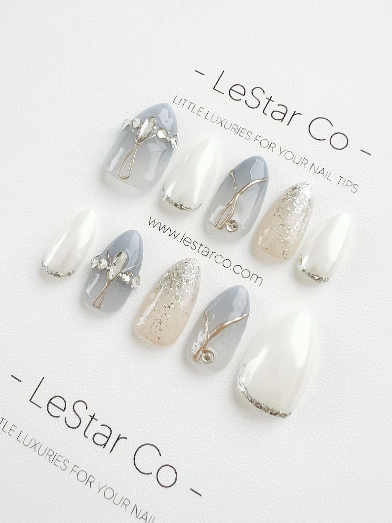 Pode incluir: Conjunto de dez unhas posti&ccedil;as com um design branco, cinza e prata com glitter. As unhas apresentam um detalhe de glitter prata, um design de espiral prata e um strass prata. As unhas est&atilde;o dispostas sobre um fundo branco com o texto "LeStar Co - Little Luxuries for Your Nail Tips" e "www.lestarco.com".