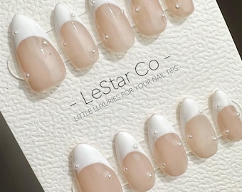 Può includere: Un set di unghie finte con un design a punta francese. Le unghie hanno una punta bianca e una base nude, con piccoli accenti di perle. Il testo "LeStar Co" e "Little Luxuries For Your Nail Tips" è stampato sulla carta. È visibile anche il sito web www.lestarco.com.