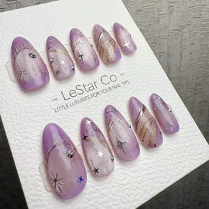May include: A set of lavender-colored nail tips with various designs. Some nails feature silver glitter, star accents, and rhinestones. Others have gold and silver swirl patterns. The text "- LeStar Co - LITTLE LUXURIES FOR YOUR NAIL TIPS" is printed on the card.