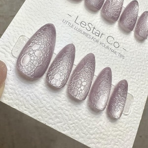 Op de afbeelding: Een set lavendelkleurige persnagels met een textuur, bubbelachtig patroon. De nagels zijn amandelvormig en gerangschikt op een witte kaart met de tekst "- LeStar Co - Little Luxuries for Your Nail Tips."