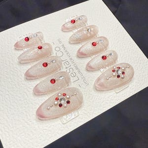 Könnte beinhalten: Set aus zehn künstlichen Nägeln mit hellrosa Glitzerbasis. Jeder Nagel ist mit roten und silbernen Strasssteinen und Perlen verziert. Die Nägel sind auf einer weißen Karte mit dem Text "Lestar Co - Little luxuries for your nail tips" angeordnet.