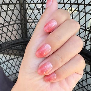 Puede incluir: Una mano con uñas ovaladas pintadas con un degradado de tonos rosa y melocotón. Las uñas tienen un acabado brillante. El fondo es un patrón geométrico negro.