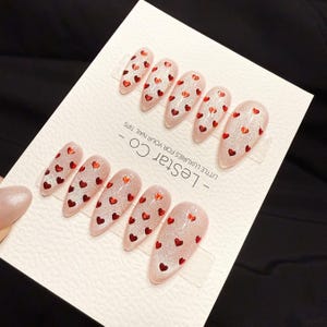 May include: Set of artificial nails with a light pink glitter finish, adorned with small red heart-shaped decorations. The nails are arranged on a white card with the text "- Lestar Co - LITTLE LUXURIES FOR YOUR NAIL TIPS".