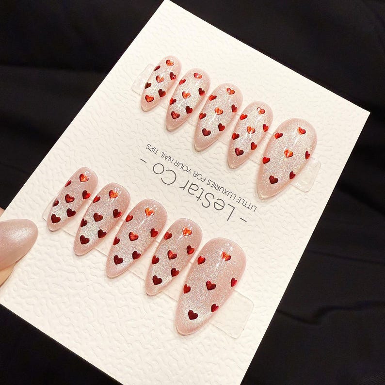 May include: Set of artificial nails with a light pink glitter finish, adorned with small red heart-shaped decorations. The nails are arranged on a white card with the text "- Lestar Co - LITTLE LUXURIES FOR YOUR NAIL TIPS".