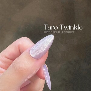 May include: A light purple nail polish with a shimmery, glittery finish. The polish has a cat eye effect, creating a line of light down the center of the nail. The text "Taro Twinkle CAT EYE EFFECT" is visible on the image.