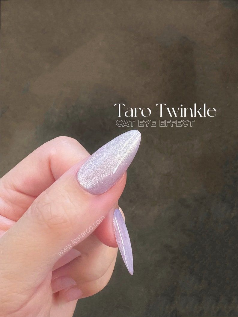 May include: A light purple nail polish with a shimmery, glittery finish. The polish has a cat eye effect, creating a line of light down the center of the nail. The text "Taro Twinkle CAT EYE EFFECT" is visible on the image.