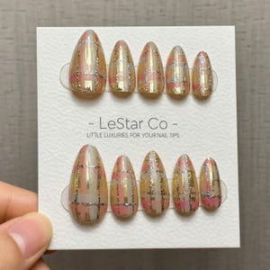 May include: A set of almond-shaped press-on nails with a plaid design. The nails feature a gold, pink, and silver color scheme. The text "- LeStar Co - LITTLE LUXURIES FOR YOUR NAIL TIPS" is printed on the card.