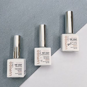 May include: Three bottles of nail polish with silver caps and white labels. The labels have the brand name "LUMIGO" and the text "NO WIPE TOP COAT" with different descriptions for each bottle: "PROTECTOR", "Enamel Diamond", and "Crystal Shine".