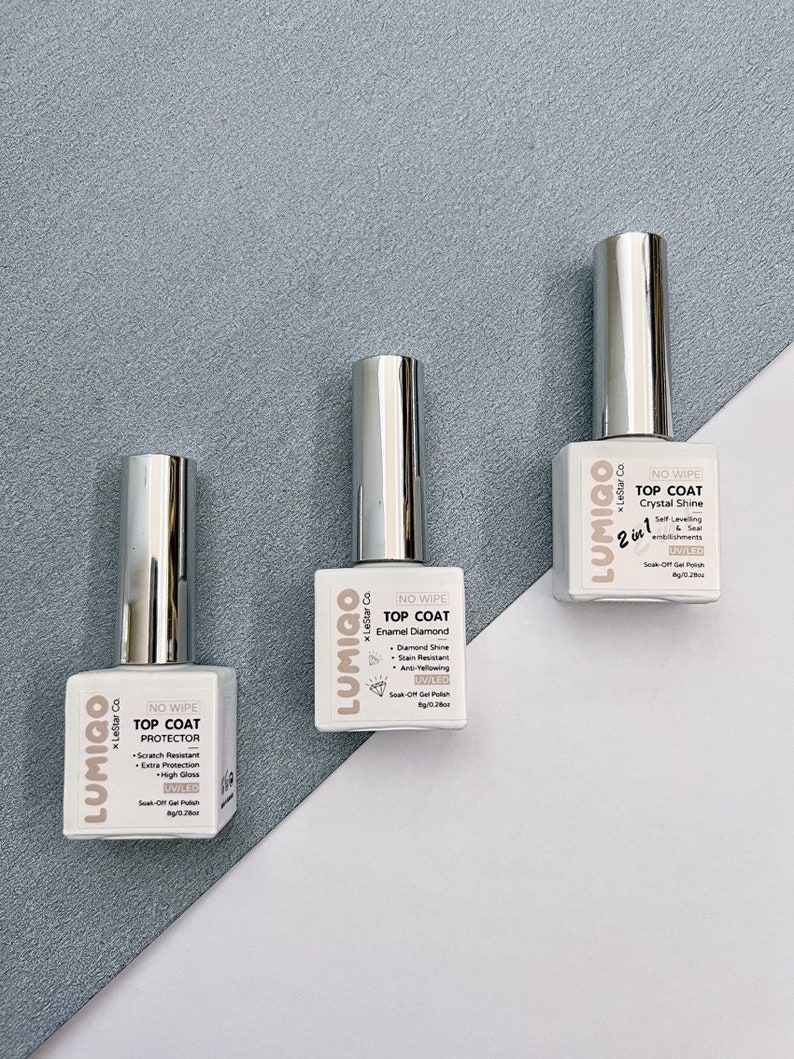 May include: Three bottles of nail polish with silver caps and white labels. The labels have the brand name "LUMIGO" and the text "NO WIPE TOP COAT" with different descriptions for each bottle: "PROTECTOR", "Enamel Diamond", and "Crystal Shine".