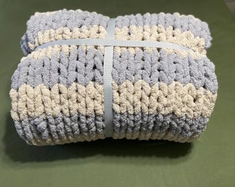Puede incluir: Una manta tejida gruesa y doblada, con rayas alternas en gris claro y crema. La manta está asegurada con una cinta gris claro. La textura del tejido es visible, lo que sugiere una sensación suave y acogedora. La manta es probablemente para la decoración del hogar.