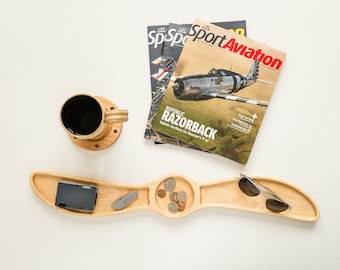 May include: A wooden tray shaped like an aeroplane propeller with three compartments. The tray has a black wallet, a small multi-tool, a pile of coins, and a pair of sunglasses.