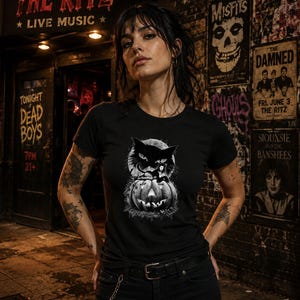 May include: A black short-sleeved t-shirt featuring a graphic print of a large black cat sitting atop a carved jack-o'-lantern pumpkin. The design is rendered in shades of black, white, and grey, showcasing a detailed illustration of the cat and pumpkin.