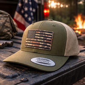Peut inclure: Casquette Snapback vert olive avec un écusson drapeau américain camouflage. Le chapeau a un dos en filet beige et une visière incurvée. Le chapeau est posé sur une surface texturée avec d'autres équipements de plein air.