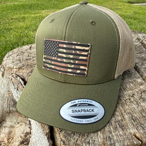 May include: An olive green snapback cap with a camouflage American flag patch. The cap has a mesh back and a curved peak. The flag patch features a black and white stars field and camouflage stripes. It has a snapback closure and a Yupoong label.