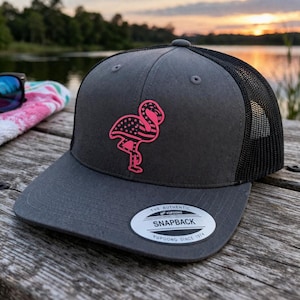Może przedstawiać: Szara czapka typu snapback z czarną siatką z tyłu, z różowym wzorem flaminga z motywem gwiazd i pasków. Czapka ma płaski daszek i białą metkę z napisem "The Authentic Snapback Yupoong Since 1974."