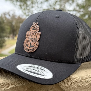 May include: Black snapback hat with a mesh back and a curved brim. The hat features a brown leather patch with the letters "USN" above an anchor design. The hat has a white tag that reads "The Authentic Snapback Yupoong Since 1974."