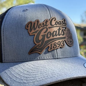 May include: A grey trucker cap with a black mesh back and a curved peak. The cap has a brown leather patch with the words "West Coast Goats 1893" in a decorative font. A USN emblem is also on the patch.