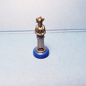 May include: A silver figurine of a person wearing a hat with horns and a long coat, standing on a blue base.