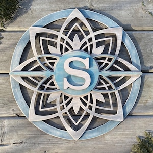 Puede incluir: Un mandala de madera con un acabado de pintura azul y blanco. El mandala tiene una gran letra "S" blanca en el centro.