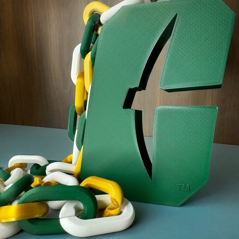 May include: A green 3D printed letter 'F' with a stylised bird design and the text 'TM' at the bottom. The letter is attached to a chain of yellow and green plastic links.
