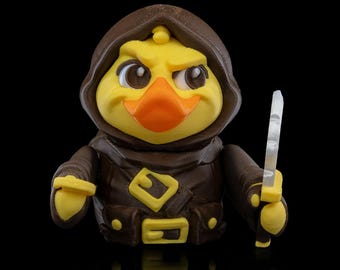 Puede incluir: Un pato amarillo con una capa marrón con capucha y sosteniendo un cuchillo plateado. El pato tiene una expresión seria y mira hacia la derecha.