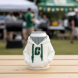 Puede incluir: Un enfriador de latas blanco con forma de sudadera con capucha, con un logotipo verde en la parte delantera. La sudadera tiene un cord&oacute;n y bolsillos. El enfriador est&aacute; sobre una superficie de madera.