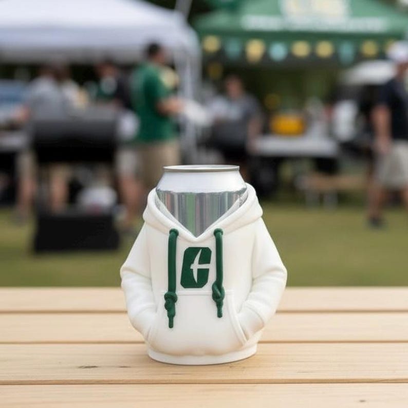 Puede incluir: Un enfriador de latas blanco con forma de sudadera con capucha, con un logotipo verde en la parte delantera. La sudadera tiene un cord&oacute;n y bolsillos. El enfriador est&aacute; sobre una superficie de madera.
