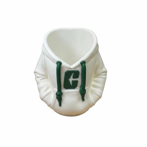 Puede incluir: Maceta de cer&aacute;mica blanca con forma de sudadera con capucha. Presenta un logotipo verde y cordones. La capucha forma la abertura de la maceta y el interior es hueco. Un art&iacute;culo decorativo.