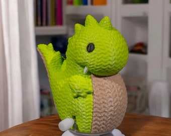 May include: A green and brown wind-up toy dinosaur with a white base. The dinosaur has a cute face with large black eyes.