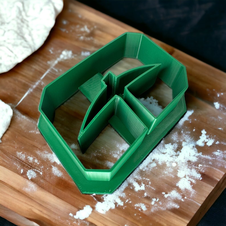 May include: A green plastic cookie cutter shaped like the letter "F" on a wooden surface covered in white flour.